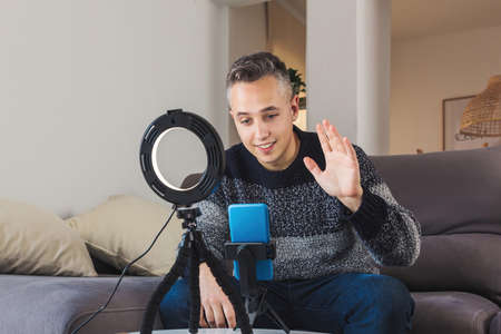Stock photo of Young man millennial influencer sit on couch is vlogging, recording and creating online content with smartphone and lights. Social media video channel recording conceptの写真素材