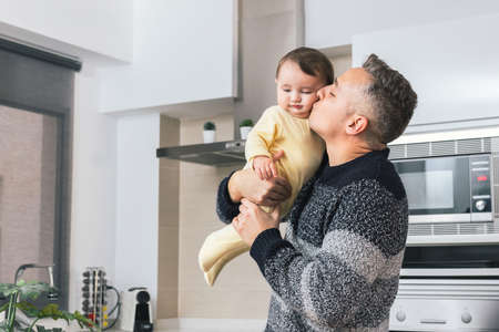 Stock photo of Young single father kissing his little baby while holding him in arms. Man and child, parent hugging child tender moment. Single dad family home life conceptの写真素材