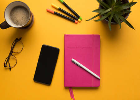 Stock photo of a pink diary with pencil, mobile phone and some desktop objects on a yellow backgroundの写真素材