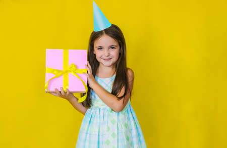 The child has a happy face holding a gift box on a yellow background. The girl has long hair, a charming happy face celebrating her birthday. The kid is happy, loves birthday and holidays.の写真素材
