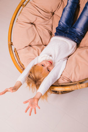 A little girl is sitting upside down in a chair. Pampering and childish jokes.Blonde girl in a white sweater.の写真素材
