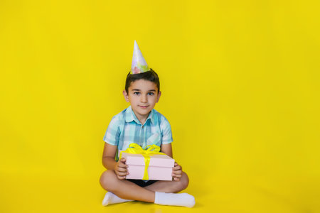A little boy sits on the floor and holds a gift on a yellow background.A birthday present.the concept of celebration and fun.a cap on the boy's head.の写真素材