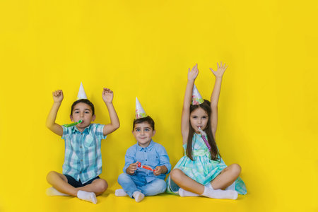 A little girl and two boys are sitting on the floor in holiday hats. Children blow their pipes and raise their hands in the air. The concept of holiday and Birthday.の写真素材