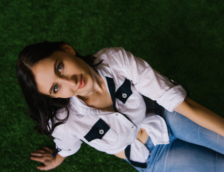 A young woman is sitting on artificial grass in a white shirt. The girl looks at the camera and...