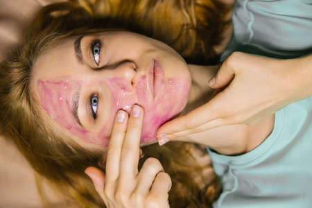 A young woman applies a pink cosmetic mask or gel to her face. Facial skin care.の写真素材