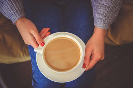 A white cup of coffee in the girl's hands. A cup and saucer are in the lap of a young woman. The woman is dressed in blue jeans and a sweater. Cozy and atmospheric photo. Winter warm drink.の写真素材