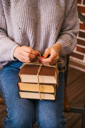 A stack of books lies on the young woman's lap. The girl ties the books with twine. The girl is wearing a sweater and jeans. Student and schoolboy. Books from the library.Textbooks close-up.の写真素材