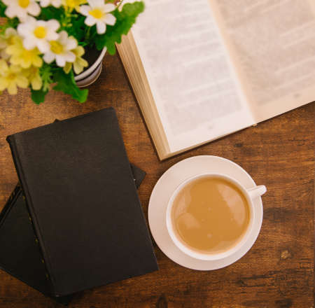 A white cup of coffee sits on a wooden table. Hardcover books lie nearby. Textbooks or fiction. The...