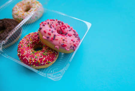 Sweet donuts of different colors in a plastic container on a blue background. Confectionery products. Home delivery of donuts. Harmful fast food.の写真素材