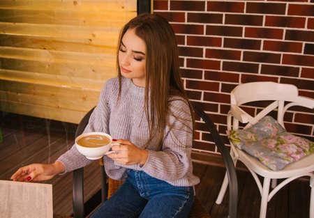 A beautiful girl in a gray knitted sweater drinks coffee and reads a book in a cozy cafe, the concept of pleasant leisure and communication. Student or businesswoman.の写真素材