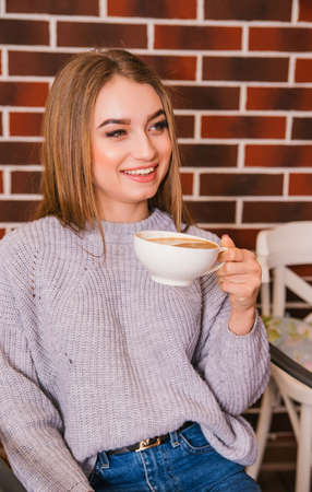A beautiful girl in a gray knitted sweater drinks coffee in a cozy cafe, the concept of pleasant leisure and communication. Student or businesswoman.の写真素材