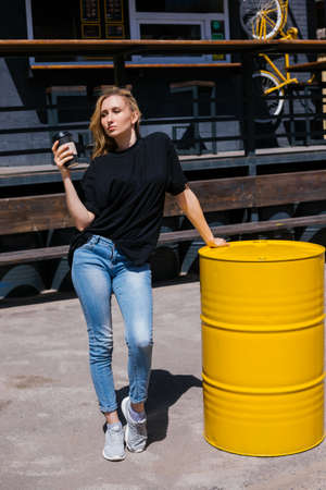 A young woman stands outside on a bright sunny day. A girl in blue jeans is holding a paper cup of...