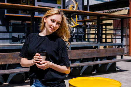 A young woman stands outside on a bright sunny day. A girl in blue jeans is holding a paper cup of coffee. Walking around the city, a way of life.の写真素材