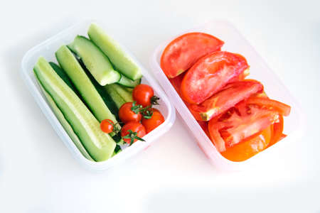 Sliced vegetables in plastic containers on a white background. Cucumbers and tomatoes are in...