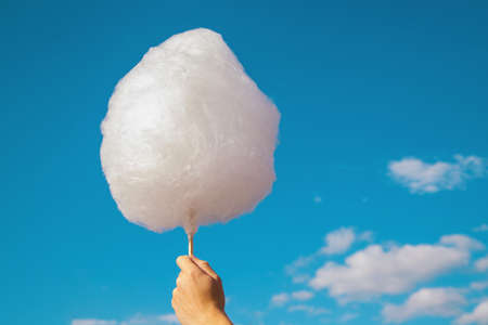 A man's hand holds cotton candy against a blue sky. White cotton wool like a cloud. A treat at the amusement park. Children's sweetness. The idea of lightness and carelessness.の写真素材