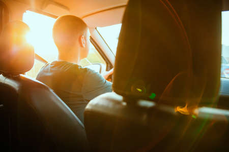A young man sits behind the wheel of a car. Outside the window is a beautiful sunset sun. The theme...