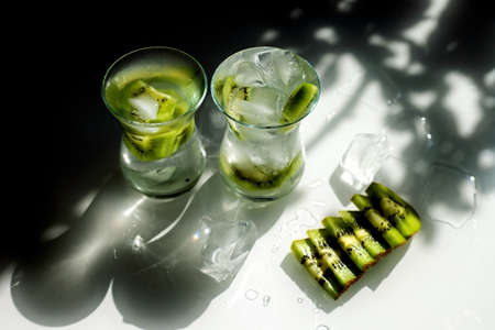 Clear lemonade with ice and kiwi slices. Two glasses stand on the table, the sun's rays falling on...