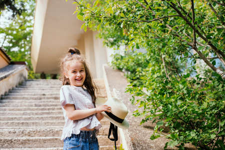 A little girl stands in the park and holds a hat in her hands. The child is happy and laughing. Summer walk in nature or in the city.の写真素材