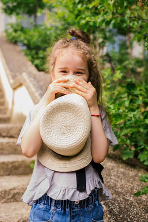 A little girl stands in the park and holds a hat in her hands. The child is happy and laughing. Summer walk in nature or in the city. The girl looks out of the hat.の写真素材