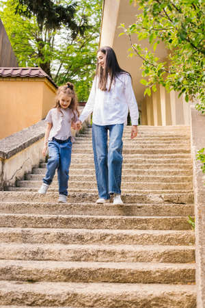 A young mother with a young daughter comes down the stairs. A woman holds a girl's hand and talks to her. Family walk in the city park.の写真素材