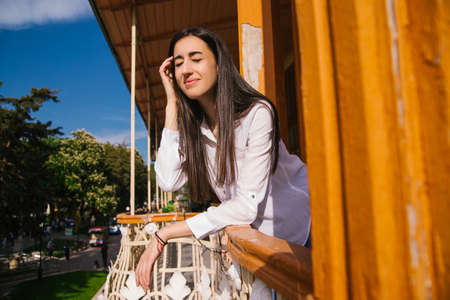 A young woman looks out from the balcony. The bright sun shines on the girl. Beautiful brunette girl enjoys the sun. The concept of recreation and travel.の写真素材