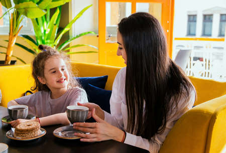 A young mother and a little daughter are sitting at the table and drinking tea. A girl and a woman sit on a yellow sofa in a cafe and have breakfast. Family leisure.の写真素材