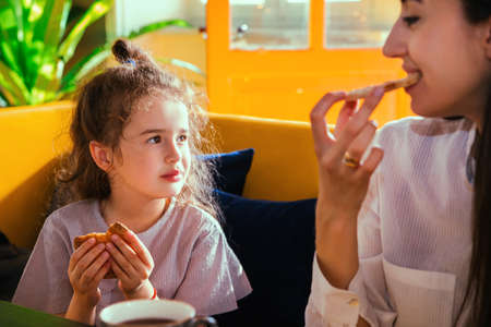 A young mother and a little daughter are sitting at the table and drinking tea. A girl and a woman sit on a yellow sofa in a cafe and have breakfast. Family leisure.の写真素材