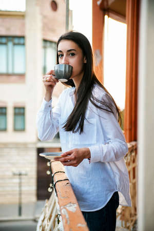 A young woman stands on the balcony and drinks coffee or tea. The girl is calm and relaxed. The concept of vacation and travel. Summer terrace or cafe.の写真素材