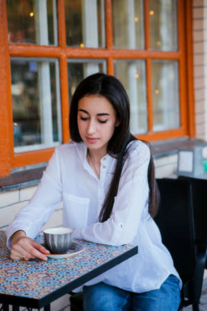 A young pretty girl is sitting at a table in a cafe and drinking coffee or tea. Rest and relaxation after a working day. A thoughtful look.の写真素材