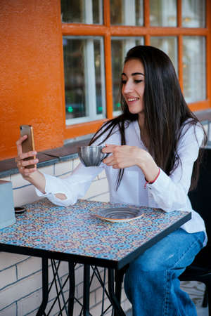 A beautiful young woman takes a selfie at a table in a cafe. A young pretty girl is talking on a video link on a mobile phone with friends or relatives.の写真素材