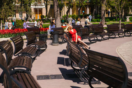A young woman sits on a bench in a city park. A girl in a red dress in the bright sun. Rest and relaxation. Spring time of the year.の写真素材