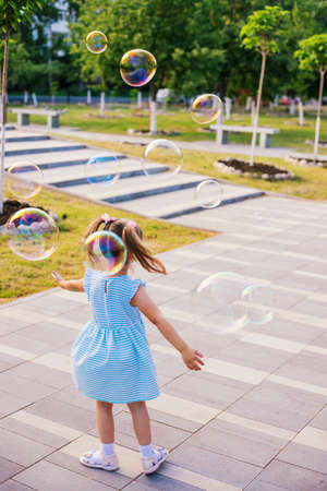 In a public park, a little girl blows and catches soap bubbles. The child is playing and laughing. The bubbles shimmer in the sun and fly in different directions.の写真素材