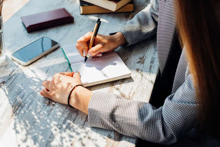 A young woman is sitting at a table by the window and writing in a notebook. The sun's rays and shadow fall on the table. A business woman works as a freelancer in a cafe.の写真素材