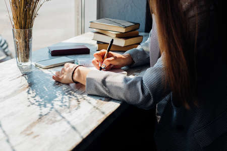 A young woman is sitting at a table by the window and writing in a notebook. The sun's rays and shadow fall on the table. A business woman works as a freelancer in a cafe.の写真素材
