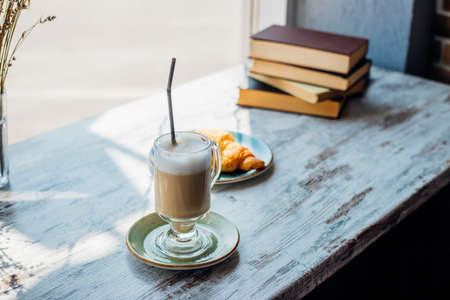 Latte in a tall glass against the background of a stack of books. The drink is on the table in a cafe, next to a croissant. Delicious breakfast and snack. Pleasant leisure and recreation.の写真素材