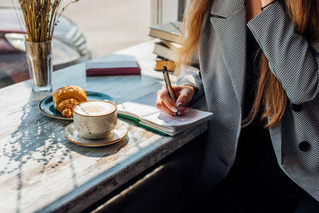 A young woman is sitting at a table by the window and writing in a notebook. The sun's rays and...