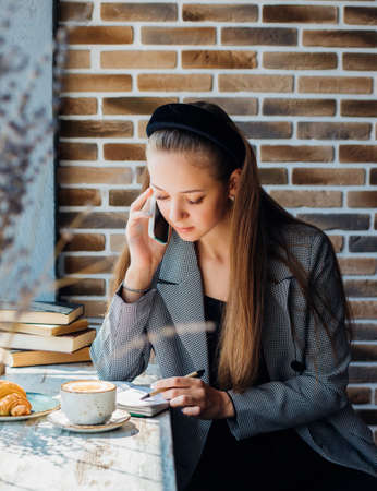 A beautiful young woman is talking on a mobile phone with friends or colleagues at a table in a cafe. The girl writes down the information in a notebook.の写真素材