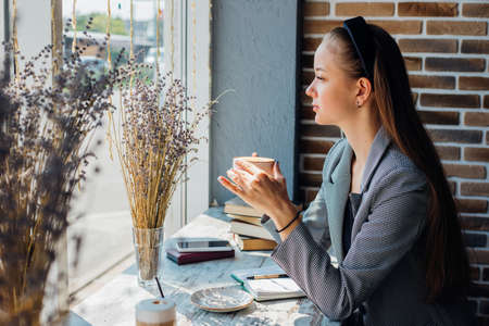 A young woman is sitting at a table by the window in a cafe and drinking coffee. The sun's rays and shadow fall on the table. The girl enjoys peace and quiet.の写真素材