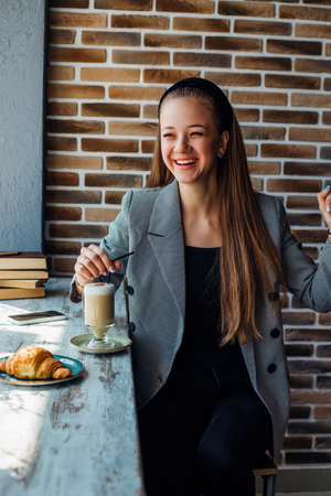 A young woman is sitting at a table by the window in a cafe and drinking coffee. The girl is laughing and dancing, in a good mood. A business woman works as a freelancer in a cafe.の写真素材