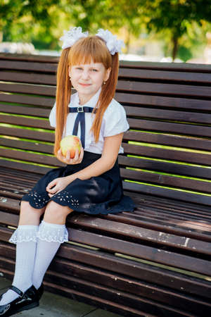Back to school. A cute little schoolgirl is sitting on a bench in the school yard and holding a green apple in her hands. Proper school meals for lunch. A little girl is going to the first grade.の写真素材