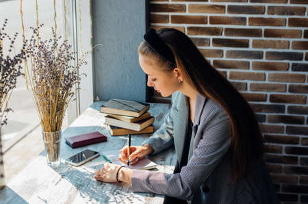A young woman is sitting at a table by the window and writing in a notebook. The sun's rays and shadow fall on the table. A business woman works as a freelancer in a cafe.の写真素材