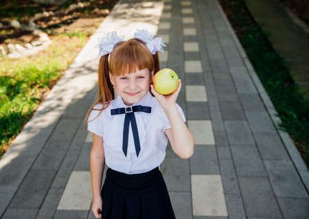Back to school. A cute little schoolgirl is standing in the park or in the school yard and holding a green apple in her hands. Proper school meals for lunch. A little girl is going to the first grade.の写真素材