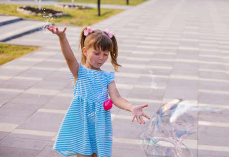 In a public park, a little girl blows and catches soap bubbles. The child is playing and laughing. The bubbles shimmer in the sun and fly in different directions.の写真素材