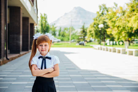 A little girl, an elementary school student, stands on the street with her hands folded. Portrait of a schoolgirl with red hair. Back to school.の写真素材
