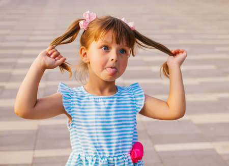 The little girl makes a face and shows her tongue. The child playfully holds his tails. Pampering a child. A fun summer walk in the park.の写真素材