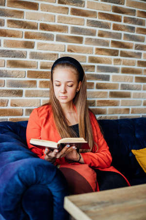 A young woman is sitting at home or in a cafe and reading a book. The girl is sitting on a soft blue sofa. The atmosphere of coziness and comfort. Pleasant leisure and self-education.の写真素材