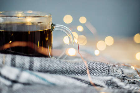 A checkered scarf with fringe lies on a light wooden background. A glass mug with black coffee stands against the background of the blurred lights of the garland.の写真素材
