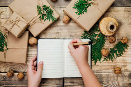 Women's hands write in a notebook with a pen. Packing boxes in kraft paper on a wooden background.Environmentally friendly material. Handmade art craft. The concept of Christmas and New Year.の写真素材
