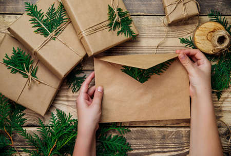 Boxes packed in kraft paper and green branches on a wooden background. Women's hands hold an envelope, a congratulatory letter. Environmentally friendly material.Christmas and New Year.の写真素材
