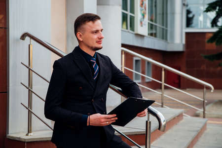 A young man in a formal suit is standing on the street with documents in his hands. managerial position. business center.の写真素材